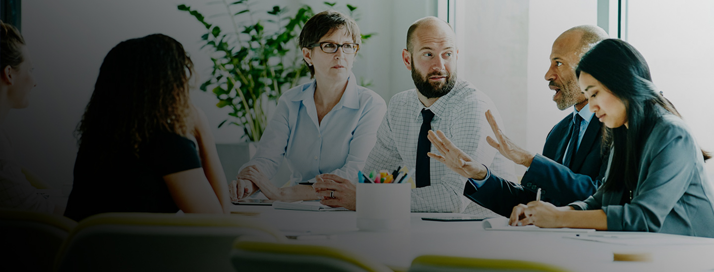 Group of people seated around a table with papers, notebooks, and coloured pencils; plant and large windows in the background.