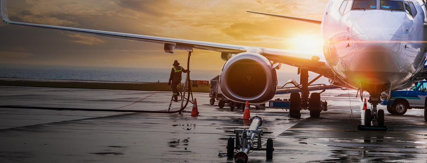 Airplane on wet tarmac at sunset with a worker in high-vis vest near the aircraft, surrounded by traffic cones.