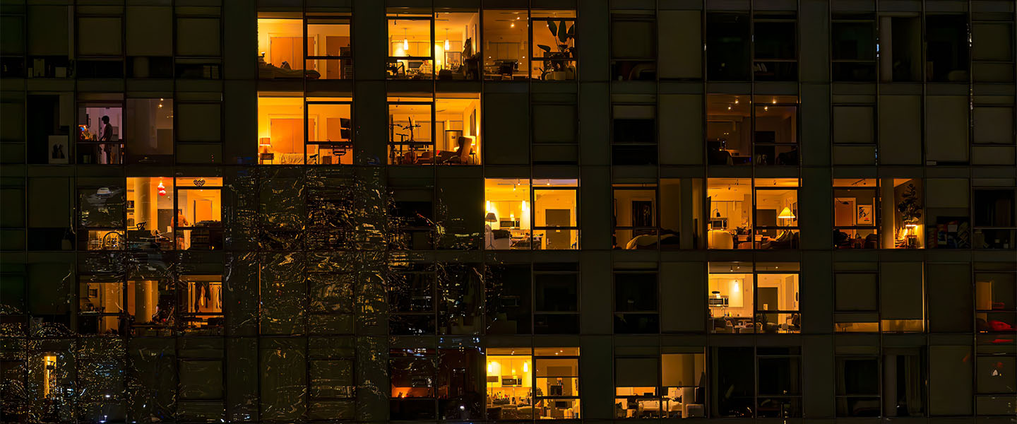 Night view of a multi-storey building with lit and dark windows showing apartment interiors.