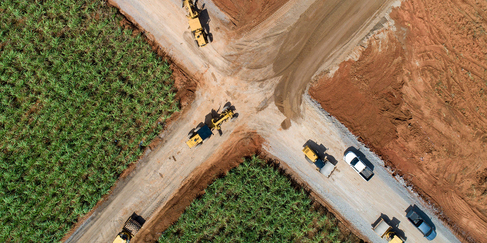 Aerial view of a construction site at a dirt road intersection, with vehicles and surrounding fields.