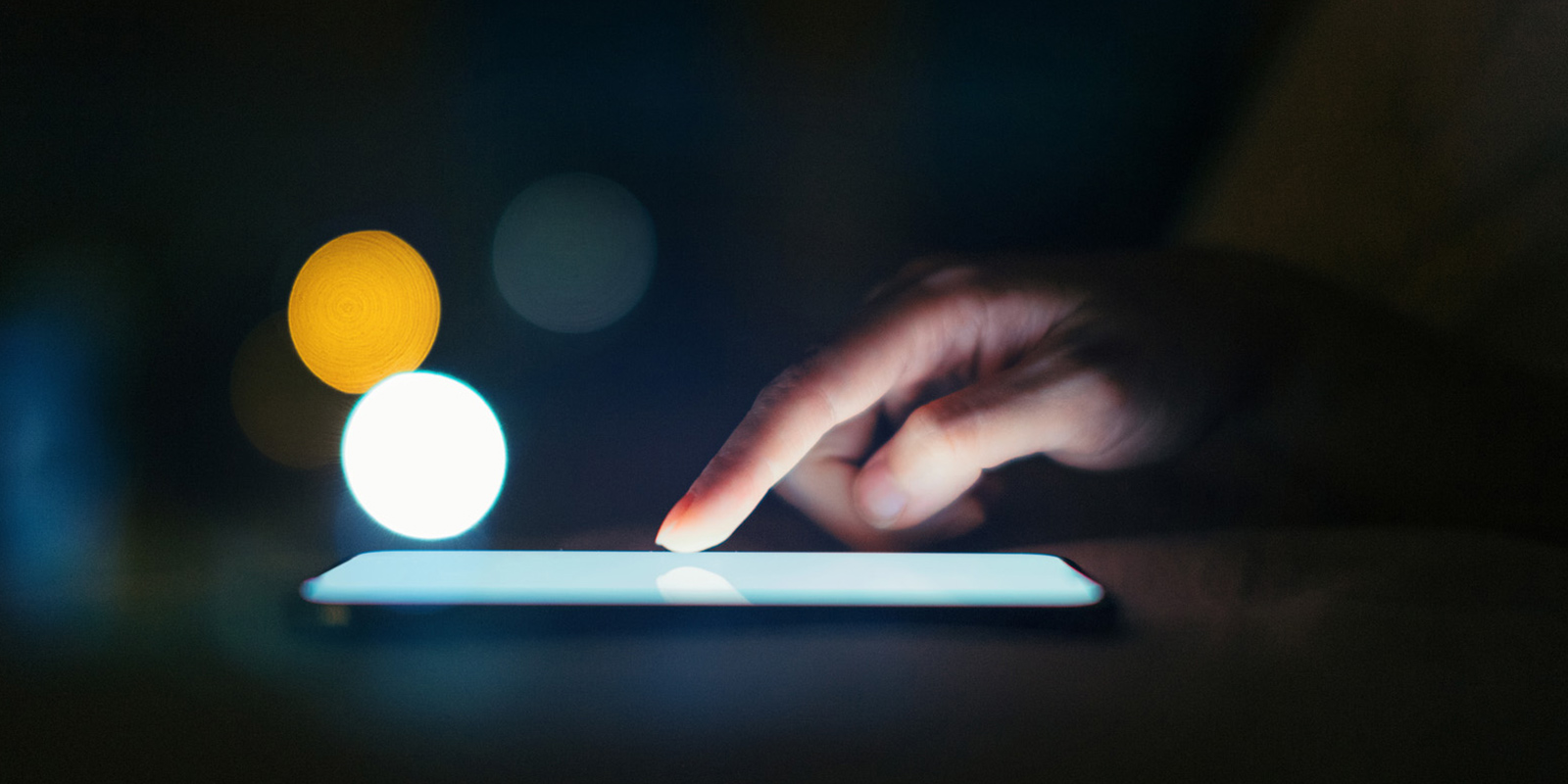 Close-up of a hand touching a lit tablet or smartphone screen with blurred lights in the background.