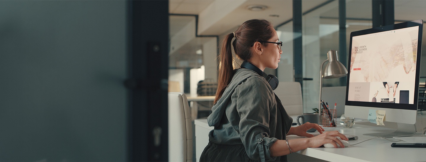 Person at a desk in an office, working on a computer with sticky notes on the monitor and stationery nearby.