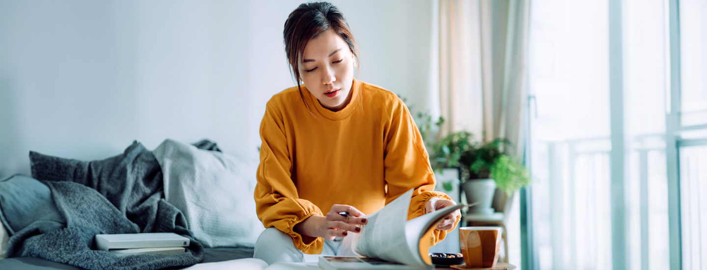 Person in yellow sweater sitting on a couch with an open book, table and cup in foreground, plants and windows behind.