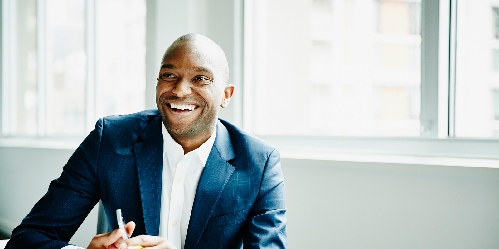 Person in a dark blue suit seated at a desk with large windows in the background.
