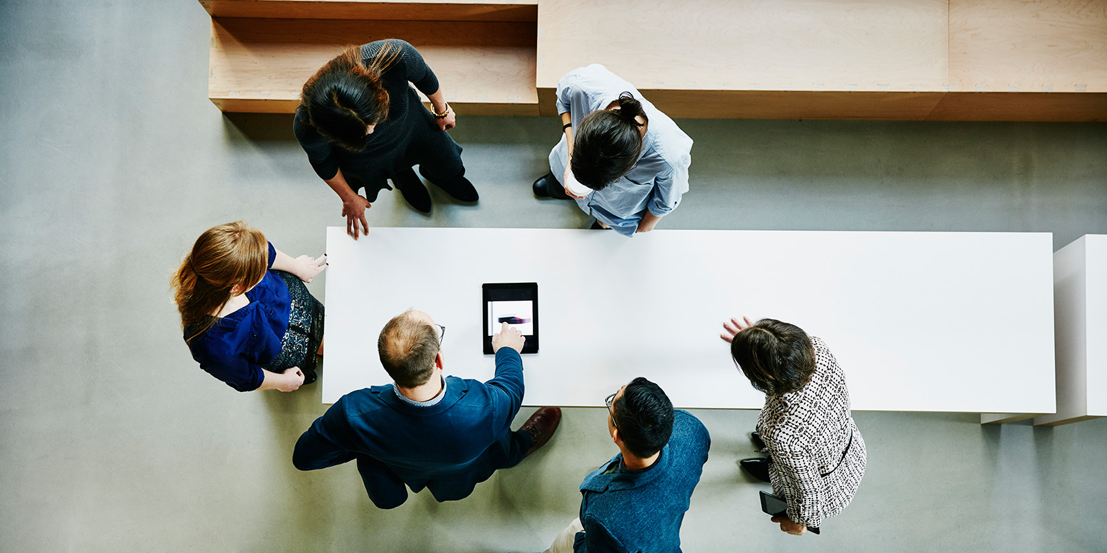 Top-down view of six people gathered around a table, with one person pointing at a tablet in the center.