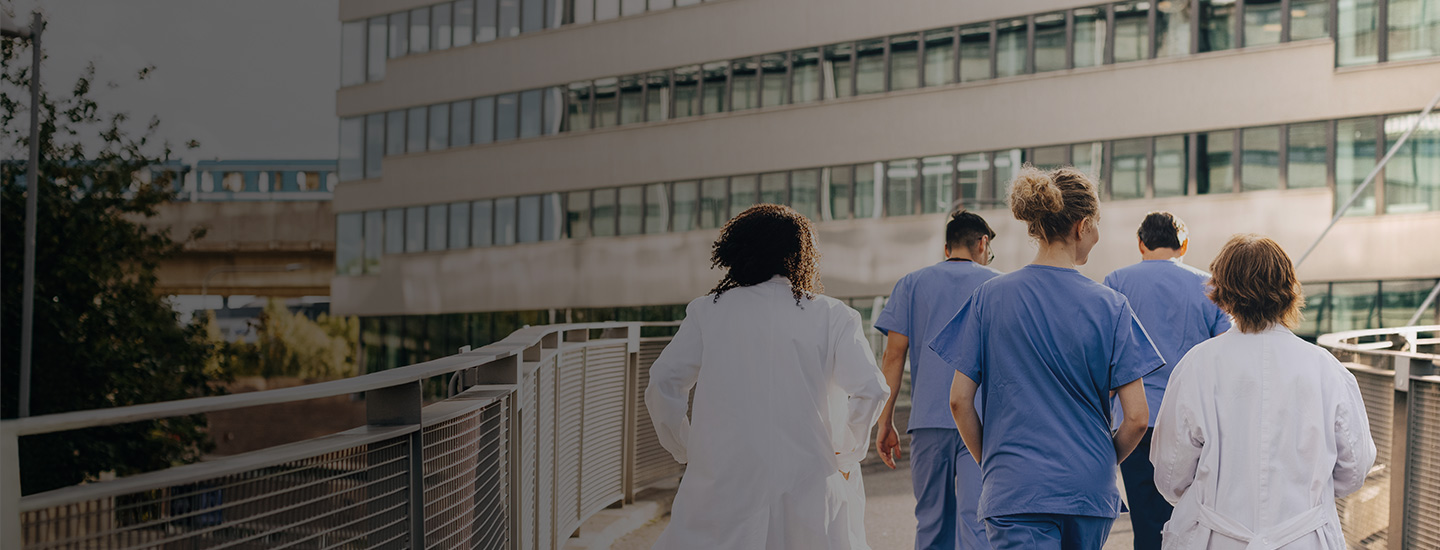 Group of healthcare professionals in medical attire walking towards a modern building with large windows.