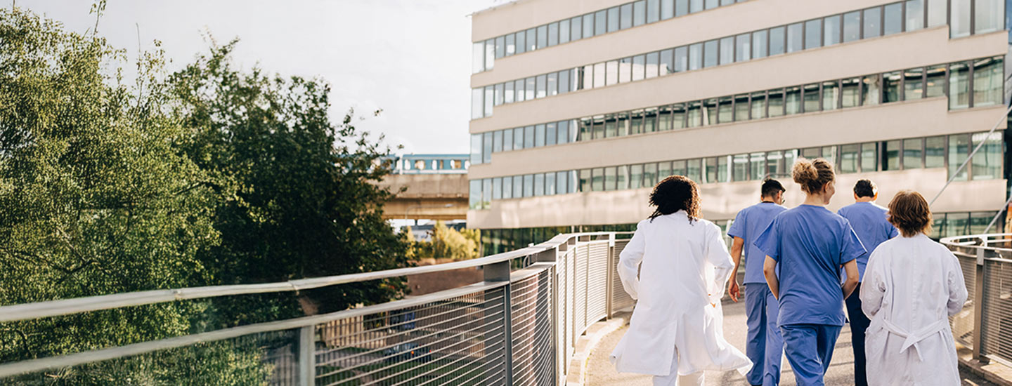 Group of medical professionals in white lab coats and blue scrubs walking along an outdoor pathway, with trees on the left.