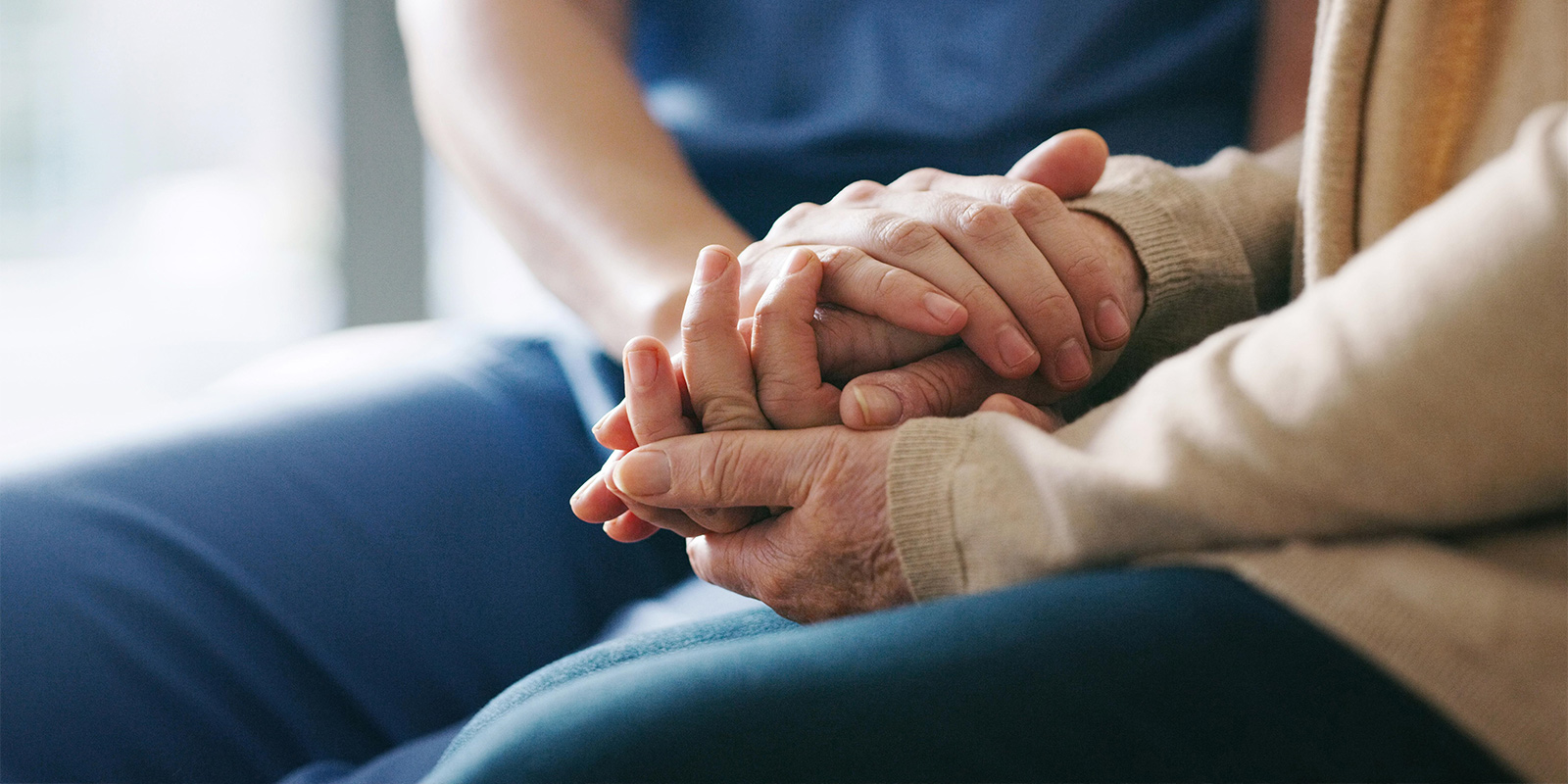 Close-up of two people holding hands, one in a blue outfit and the other in a beige sweater, suggesting comfort or support in a healthcare setting.