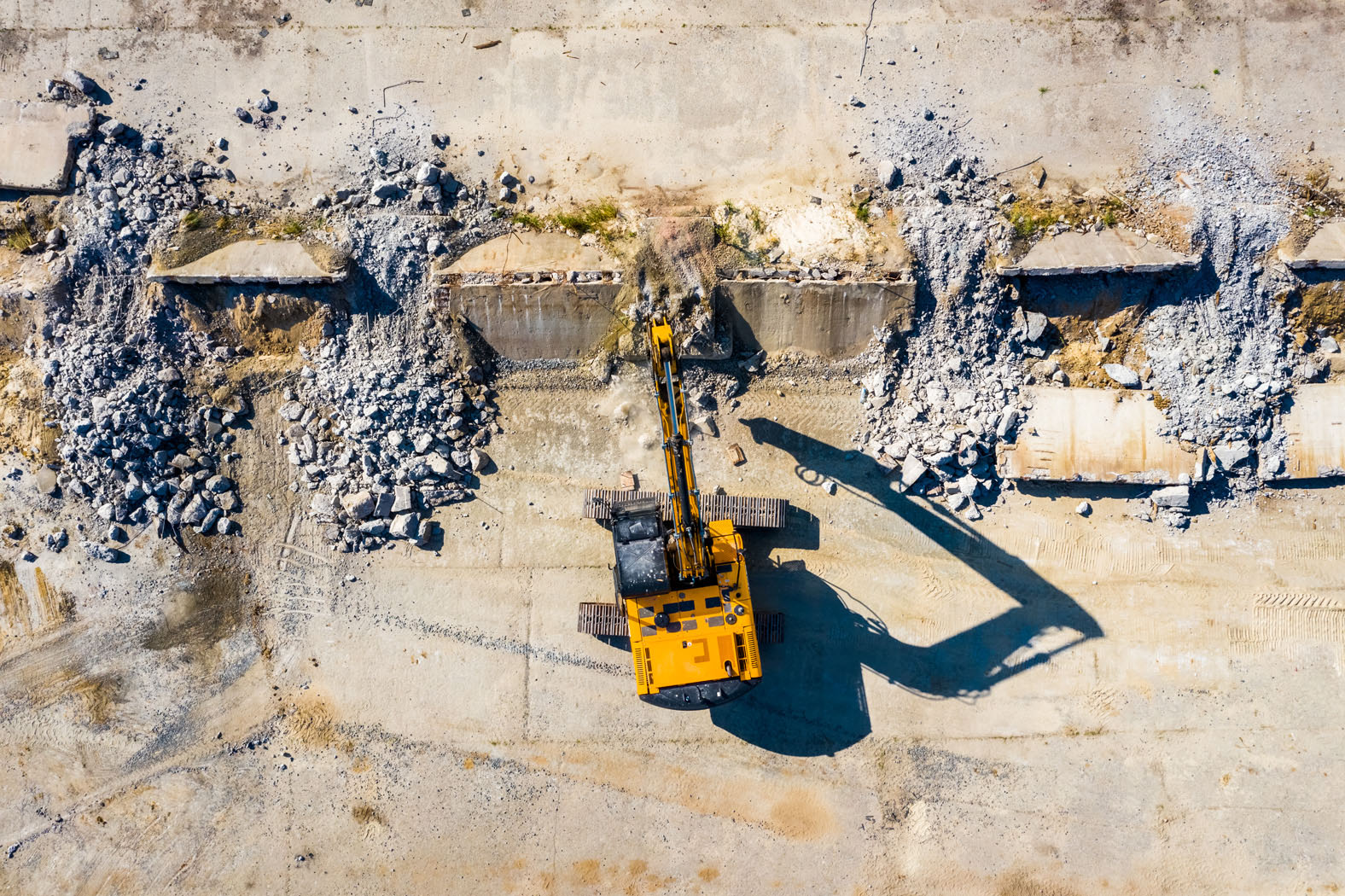 Aerial view of a construction site with a yellow excavator demolishing a concrete structure, surrounded by rubble.