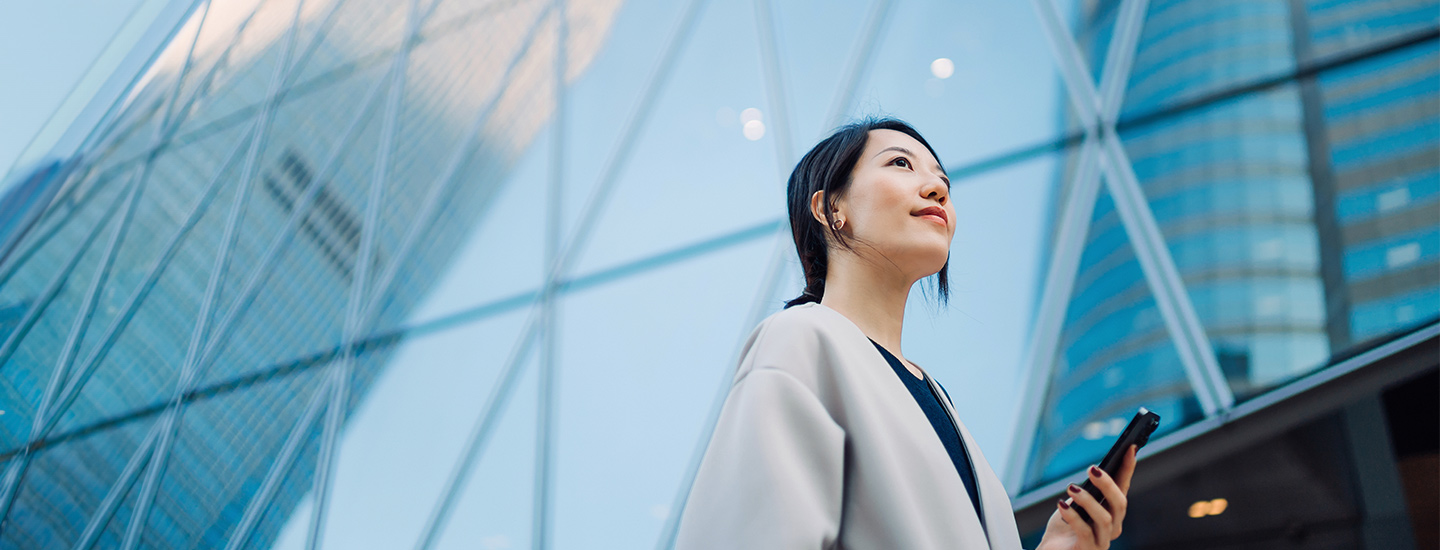 Person holding a smartphone in front of a modern glass building reflecting nearby skyscrapers.