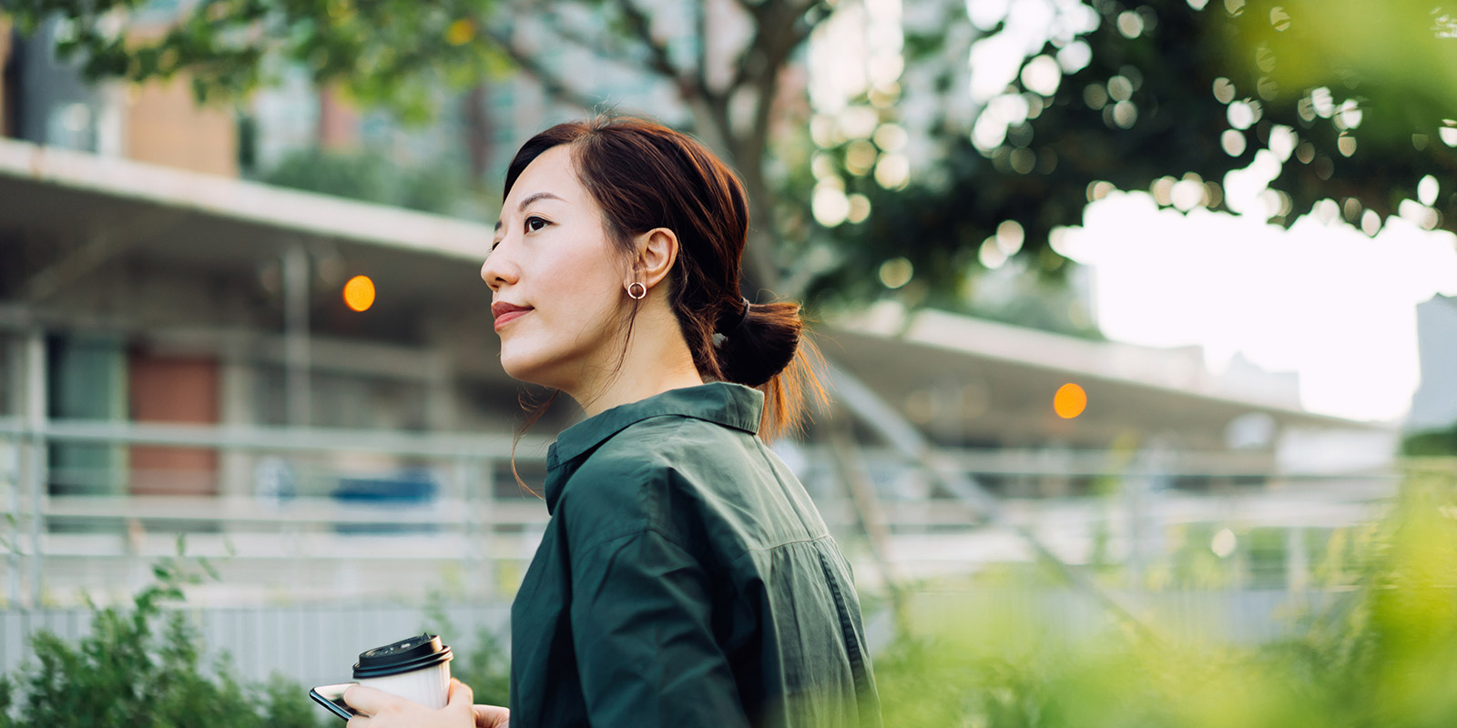 Person in a green jacket holding a cup and phone, standing outdoors with trees, greenery, and buildings in the background.