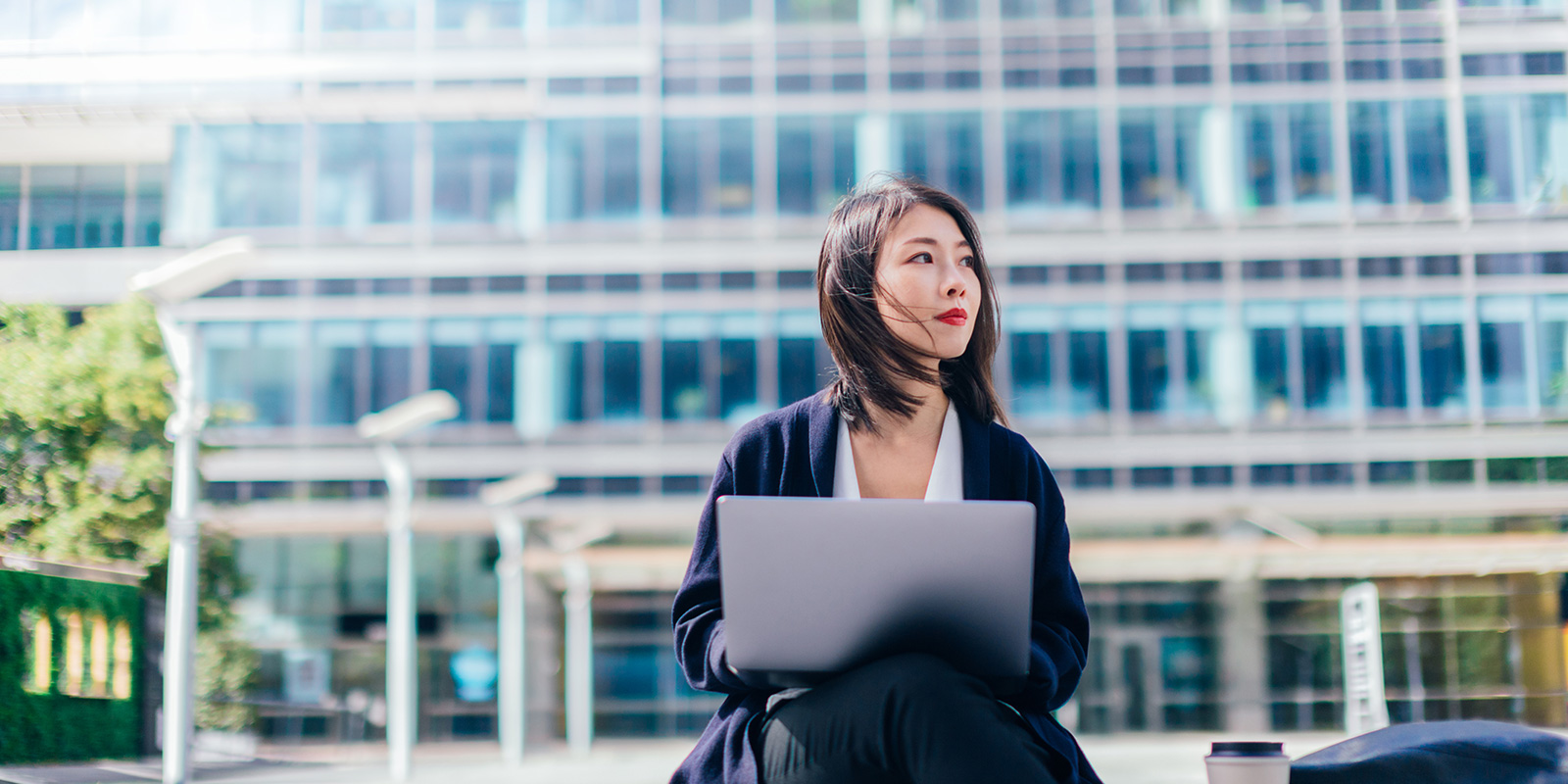 Person sitting outdoors with a laptop in front of a modern glass building.