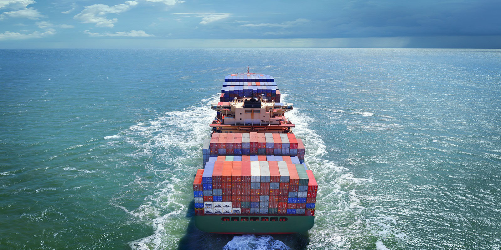 Aerial view of a large cargo ship loaded with containers, sailing through ocean water under a partly cloudy sky.