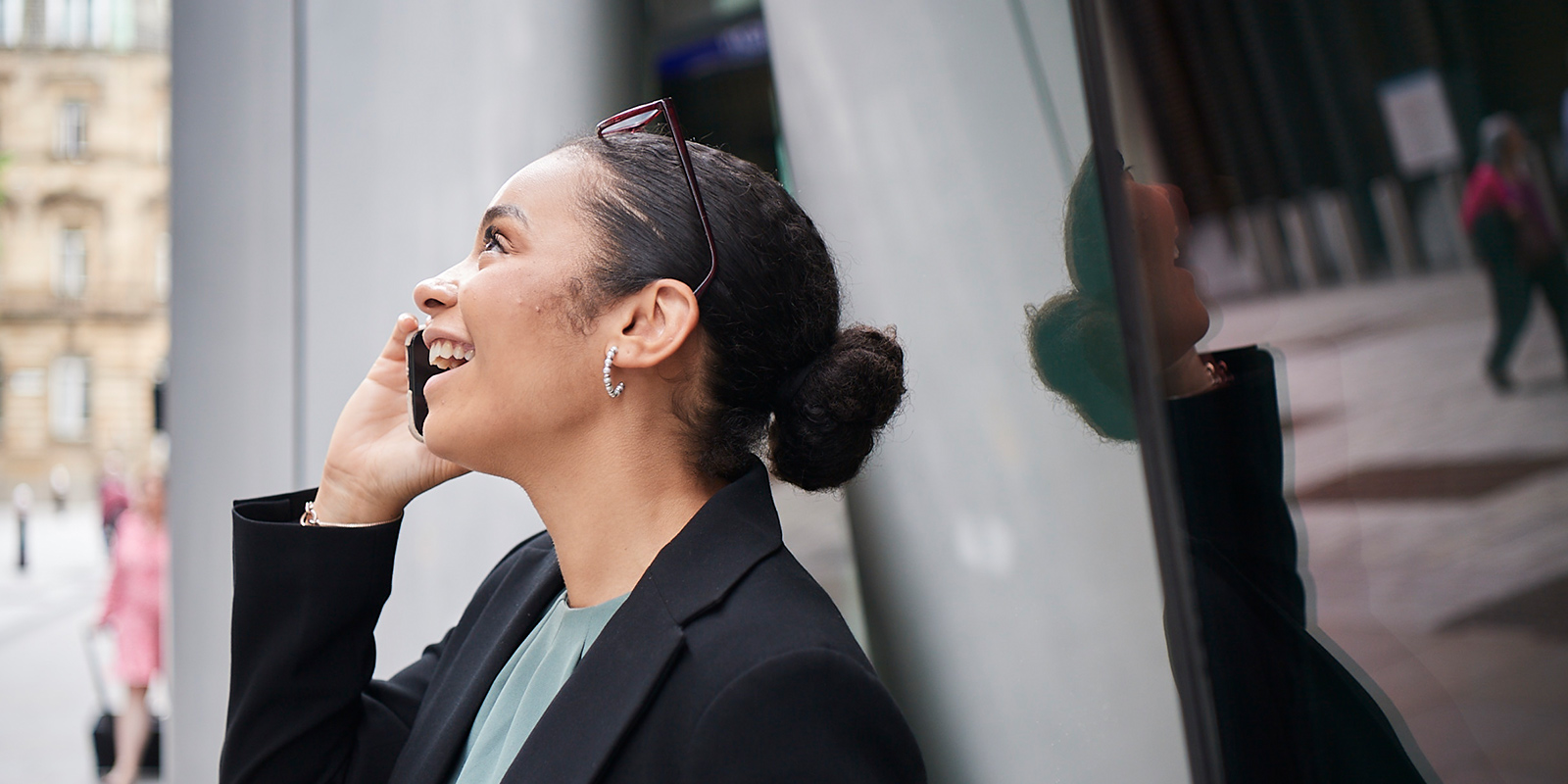 Person wearing a black blazer and holding a phone to their ear beside a reflective glass surface in an urban setting.