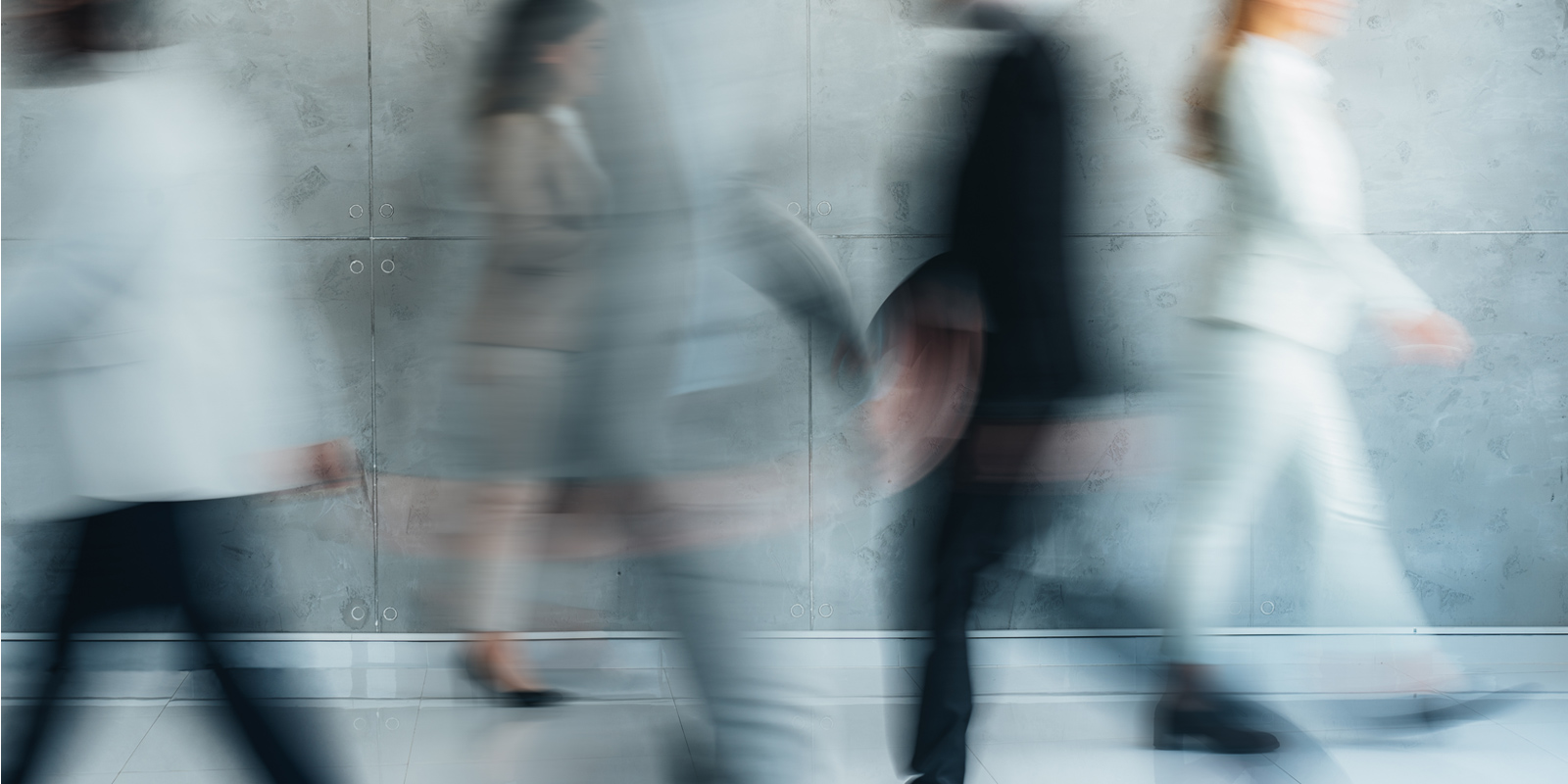 People in business attire walking past a concrete wall in blurred motion.