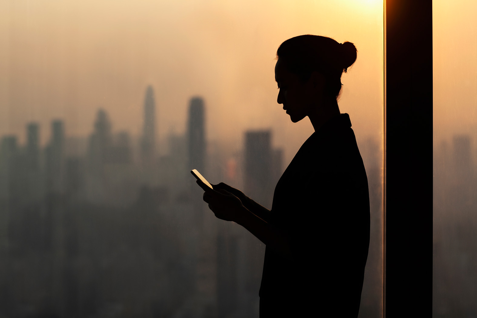 Person's silhouette by window, on phone, with city skyline in the background.
