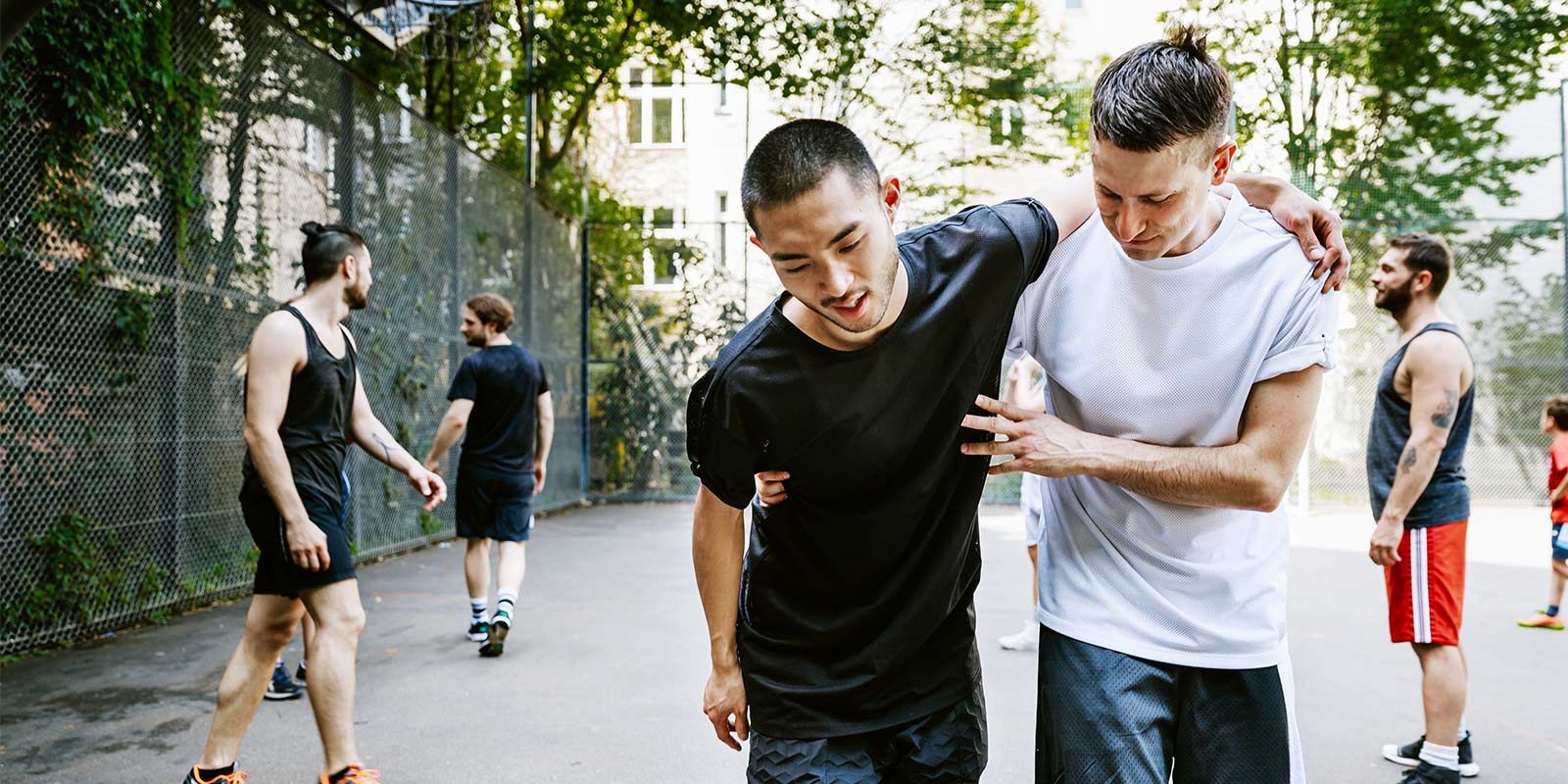 Person in white shirt helps injured player in black shirt off outdoor basketball court during a game.