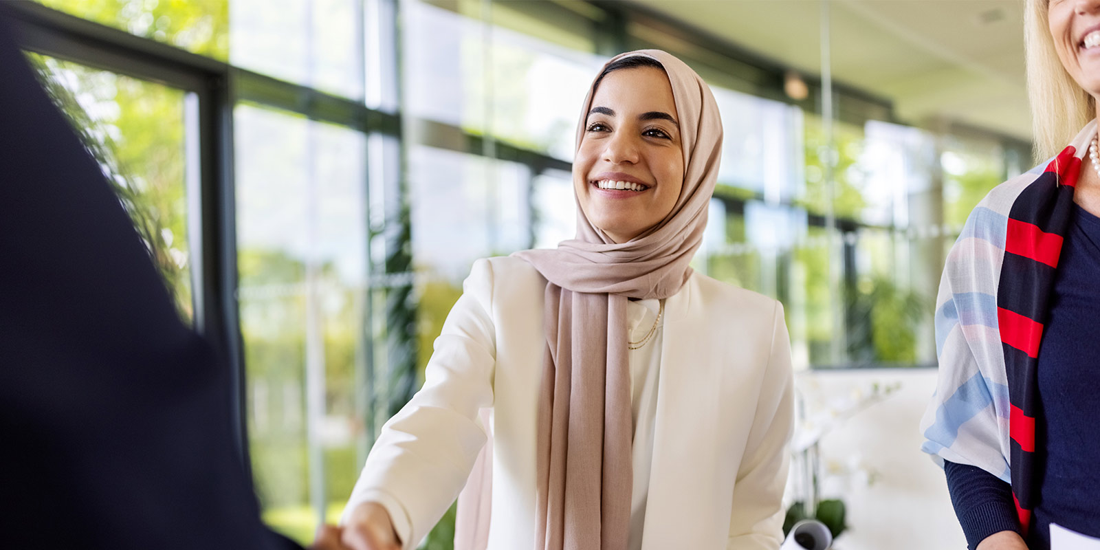 Three people in a modern office, with one wearing a white blazer and beige headscarf shaking hands, and another partially visible with a striped scarf.