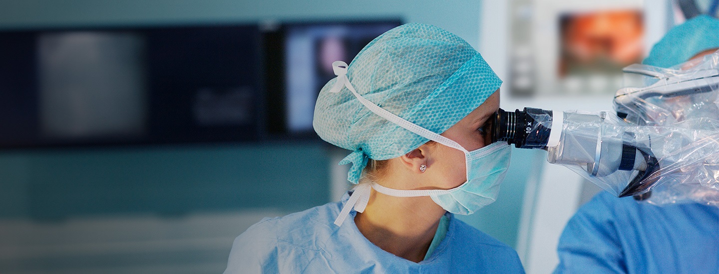 Medical professional in blue gown and cap using a microscope in an operating room with blurred equipment in the background.