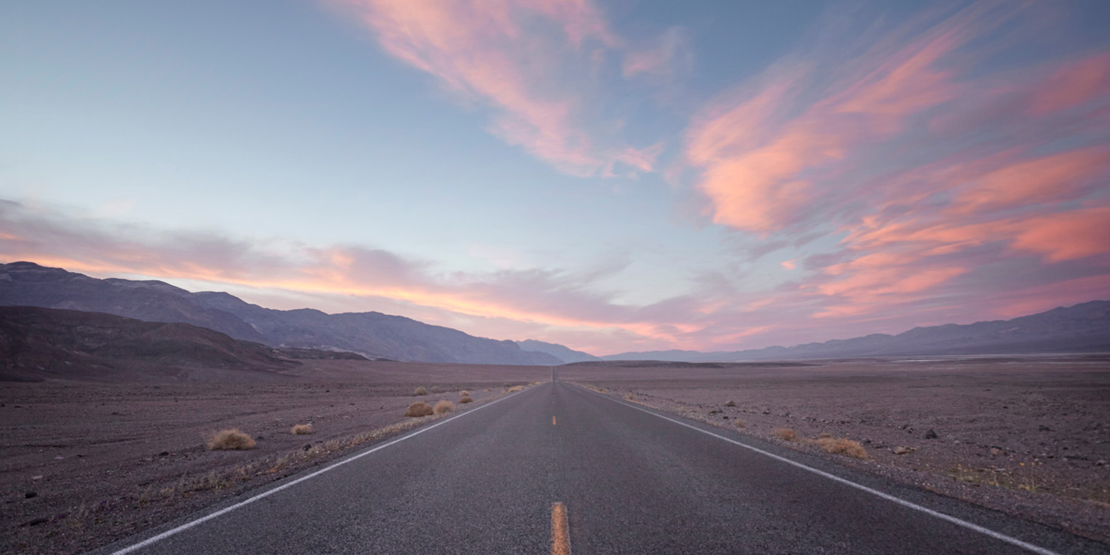 Straight road through a barren desert landscape with pink-orange clouds and distant mountains.