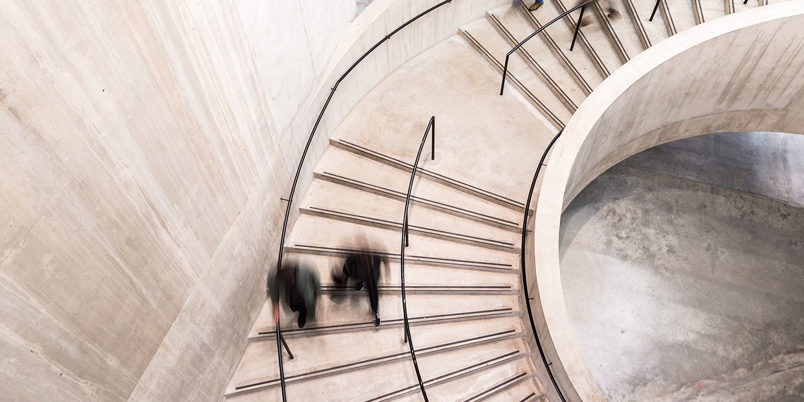 Spiral concrete staircase with metal railings and two blurred figures walking.