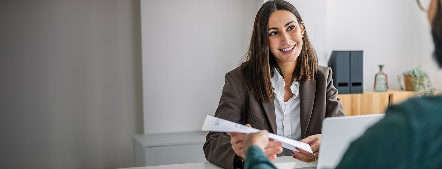 Two people seated across a desk in an office, with one passing a document to the other.