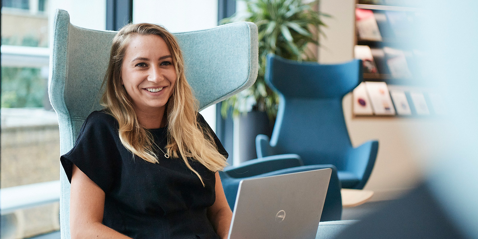 Person working on a laptop in a modern office with large windows, plants, and contemporary furniture.