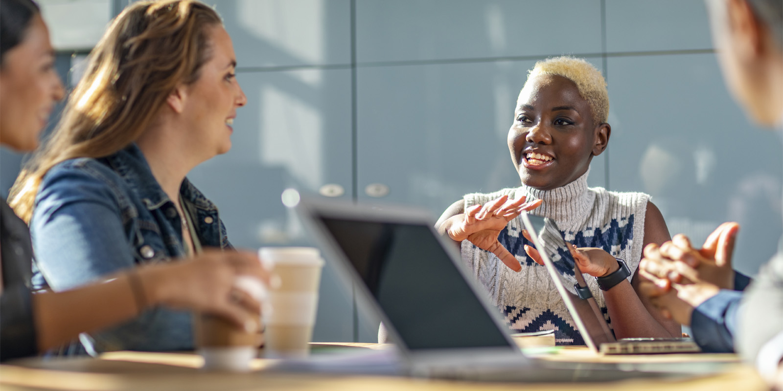 Group of people seated around a table with laptops and coffee cups, one person gesturing while speaking.
