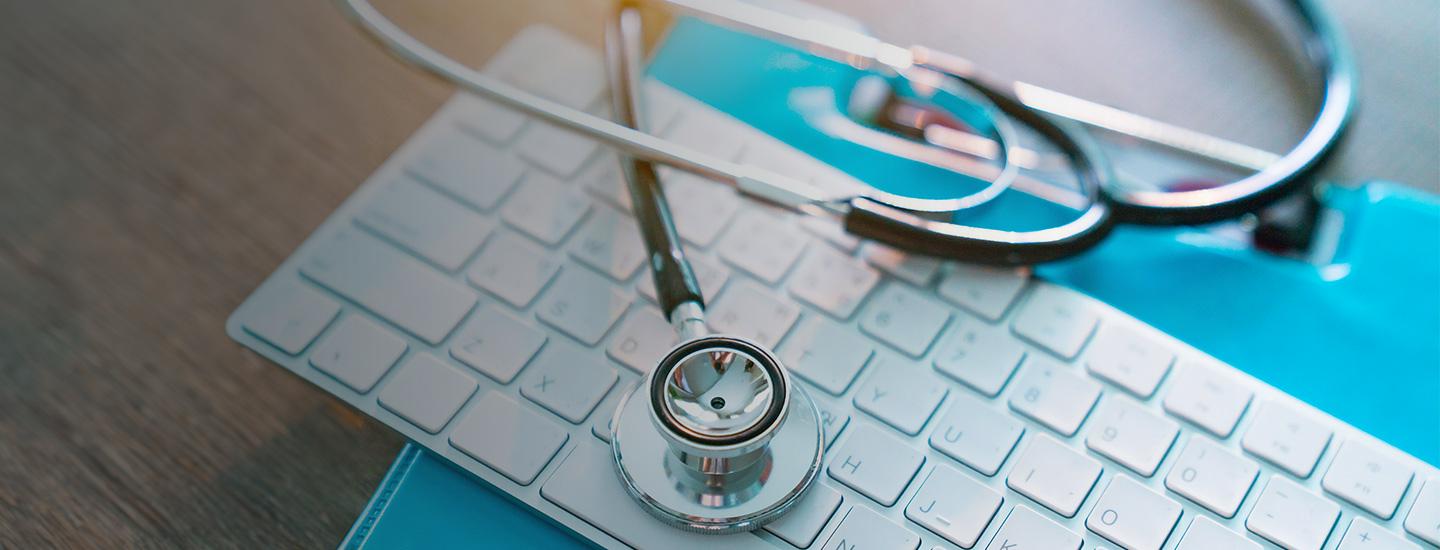 Stethoscope resting on a white computer keyboard with blue folders partially visible beneath, on a wooden surface.
