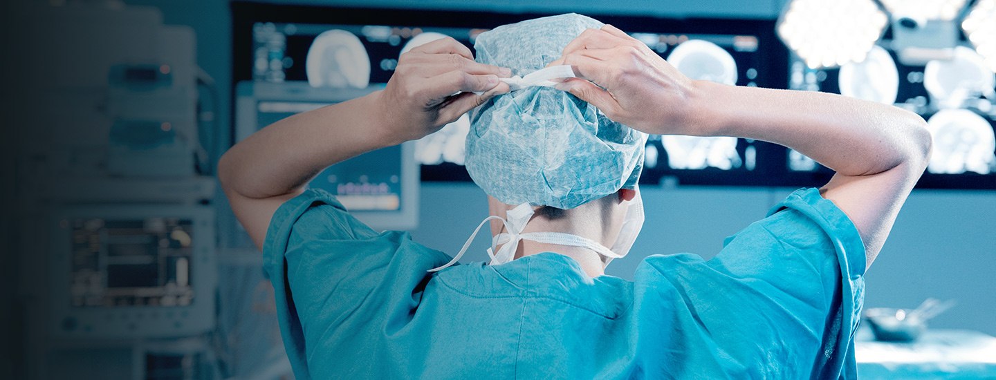 Medical professional in surgical scrubs adjusting a cap, with monitors showing medical scans in an operating room setting.
