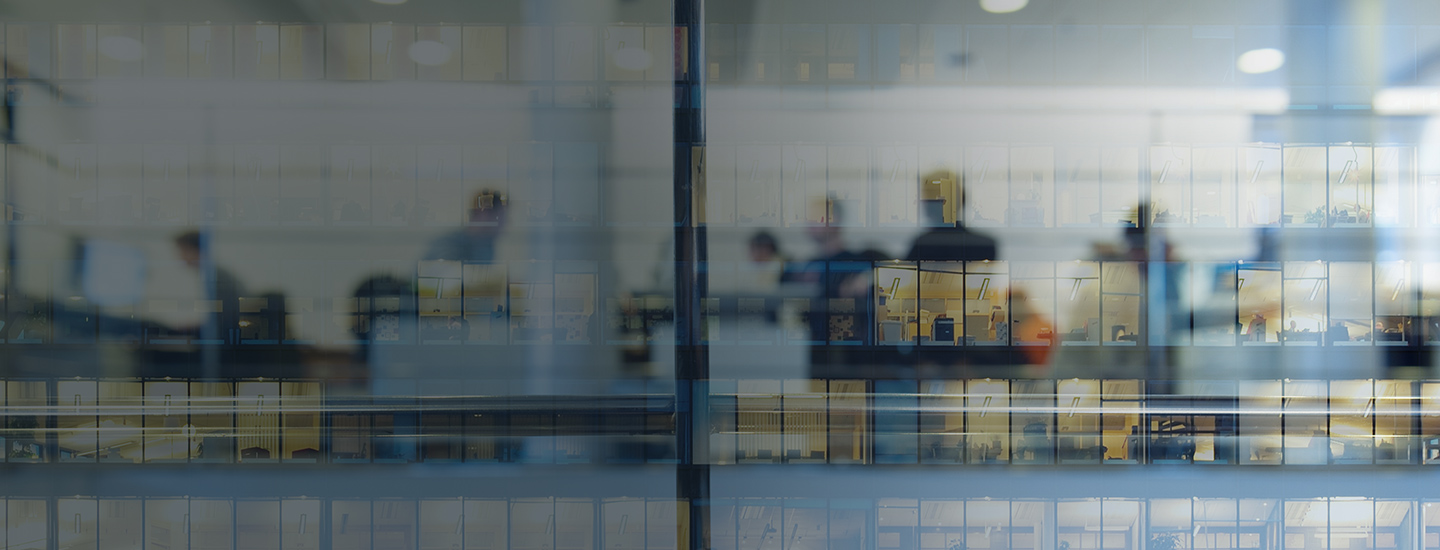 Reflection of a window showing people working inside a multi-floor office building, with desks and computers visible.