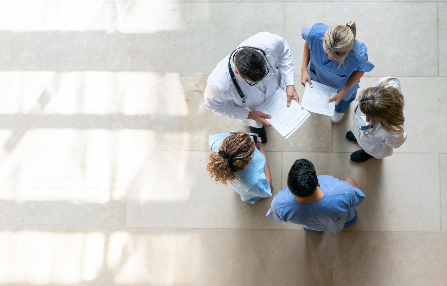 Five medical professionals in scrubs and lab coats stand in a circle indoors, viewed from above.