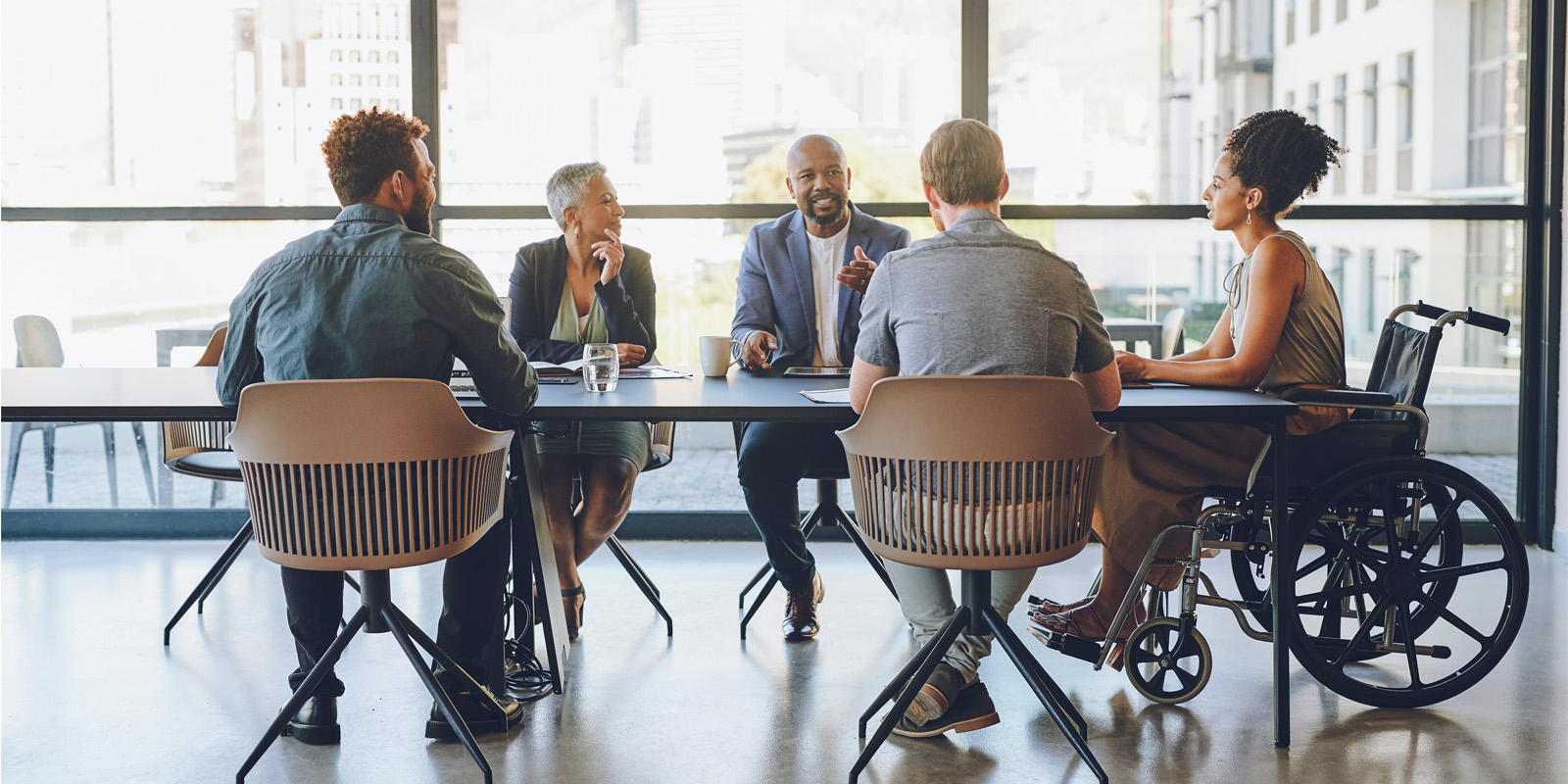 Five people in discussion around a conference table in an office, with one person using a wheelchair and city views outside.