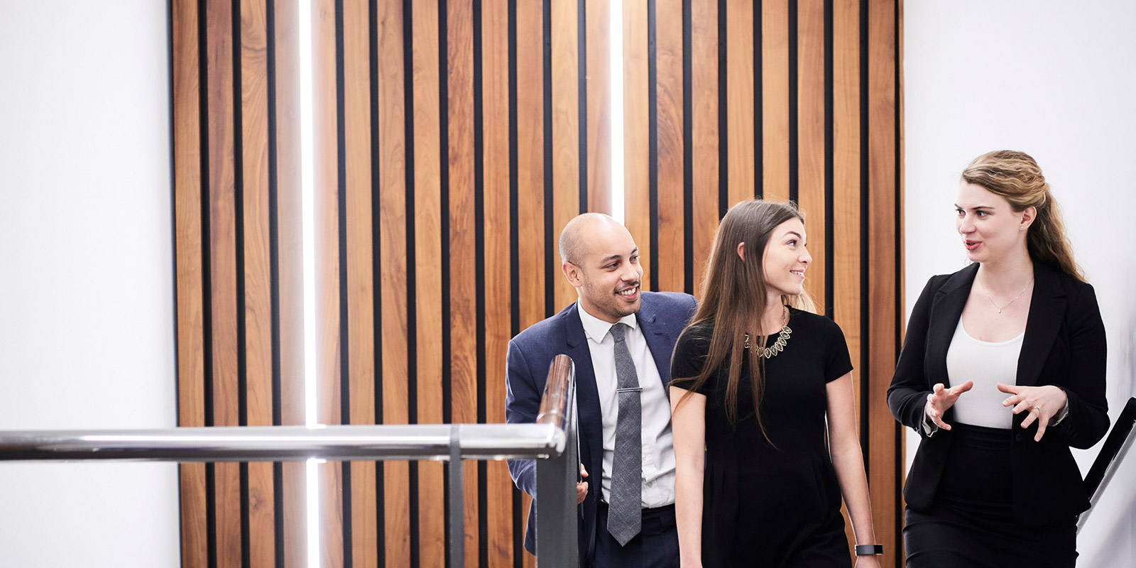 Three people in business attire walking upstairs past a wooden panel wall.