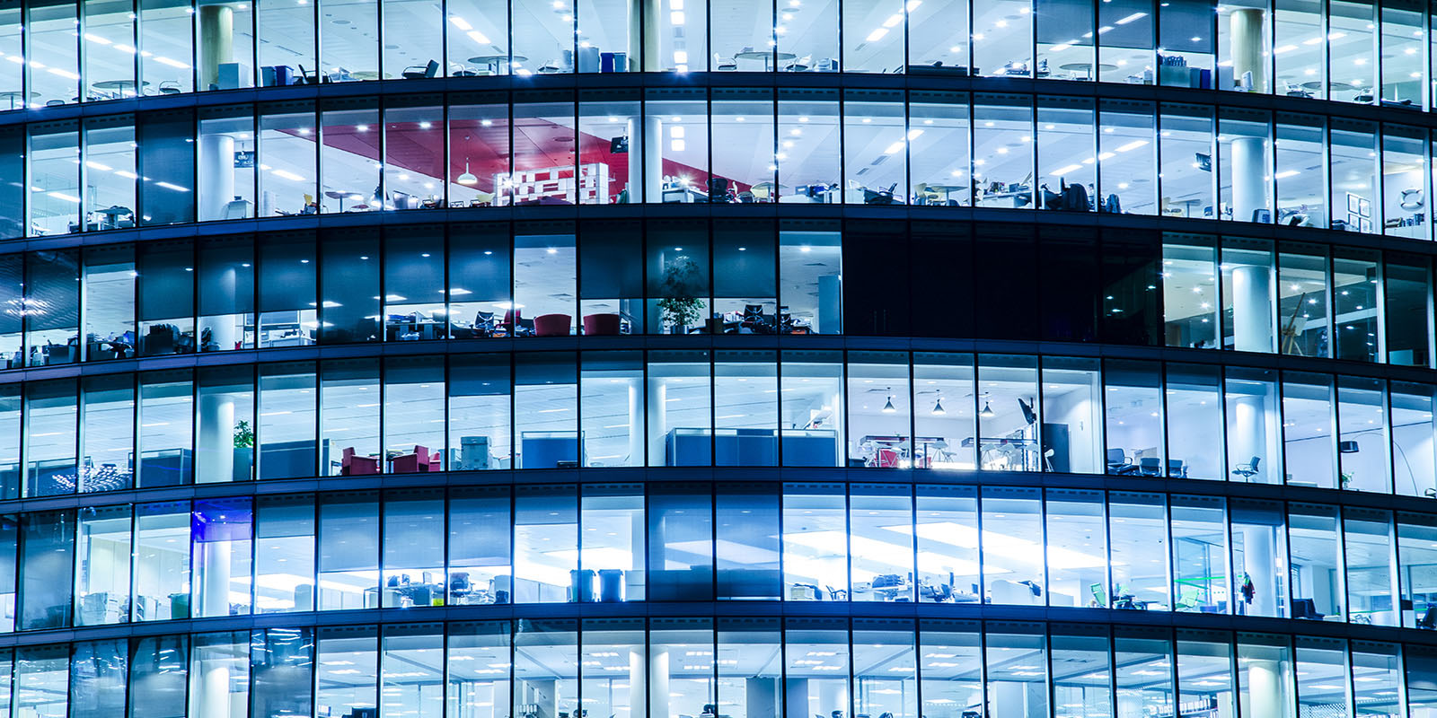 Multi-storey office building with large glass windows showing lit interiors and desks.