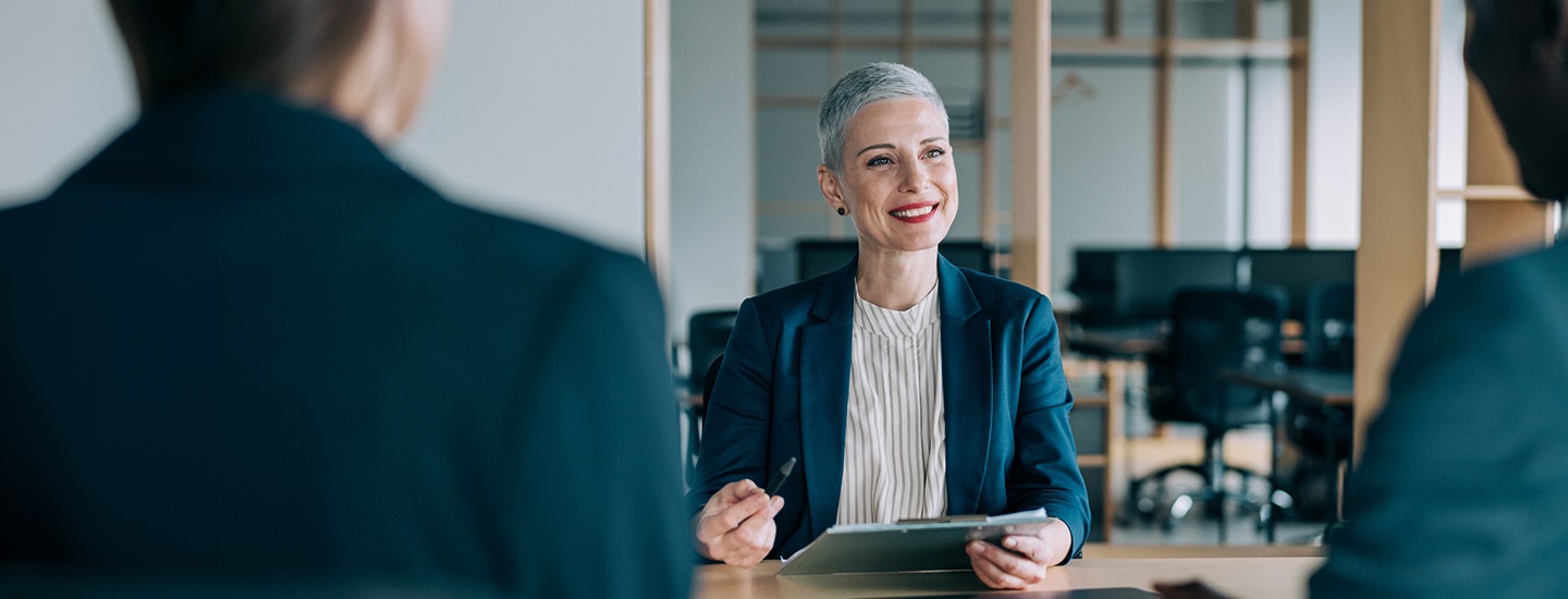 Person in a blazer writing on a clipboard at a desk, with two others partially visible in the foreground.