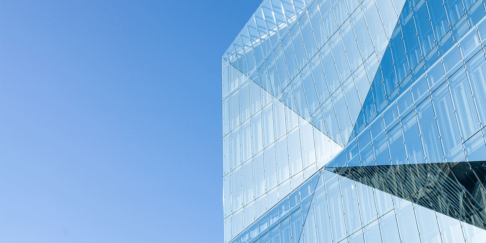 Modern glass building with sharp angles and mirrored panels reflecting a clear blue sky.