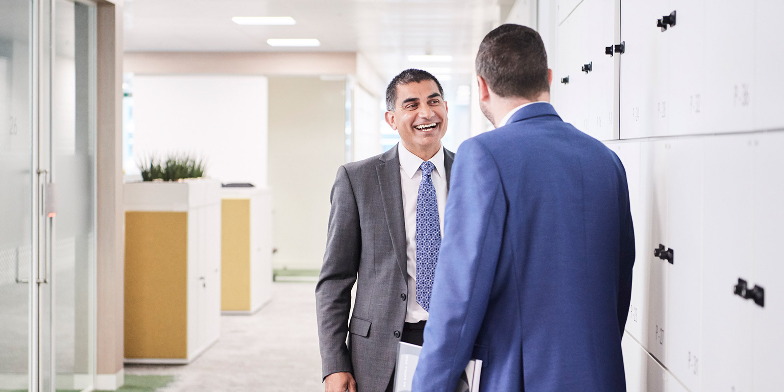 Two people in business attire talking in an office hallway with lockers and cabinets in the background.