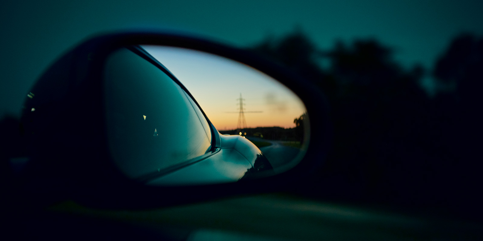 Reflection in car side mirror showing road and transmission tower at twilight.