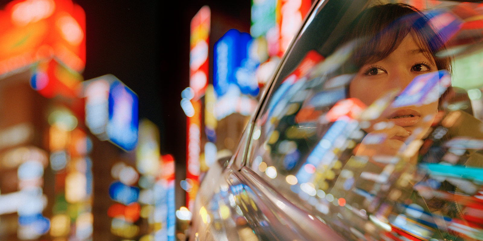 Blurred cityscape at night with neon lights reflected on a car window; a person is visible inside.
