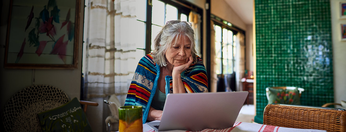 Person wearing a colourful striped shawl, seated at a table using a laptop in a bright room with green tiles.
