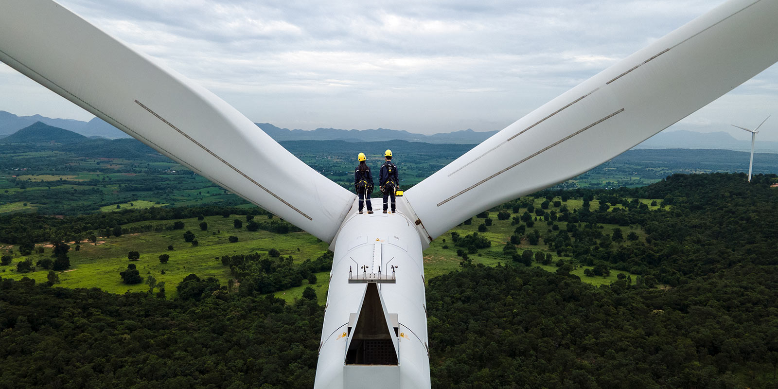 Two workers in safety gear standing on top of a wind turbine in a rural area with fields and hills in the background.