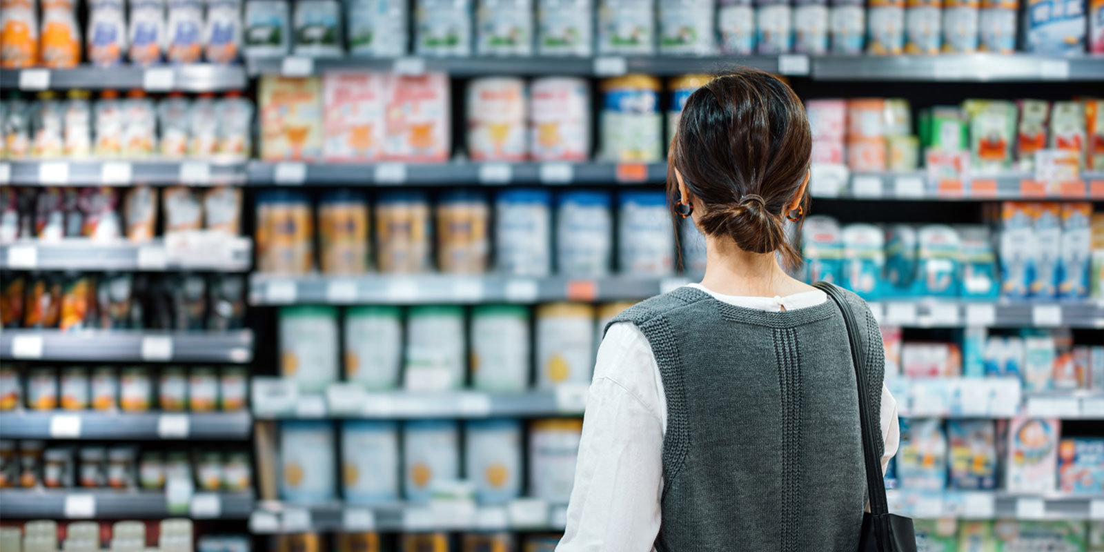 Person in grey vest browsing shelves of canned and packaged goods in a store.