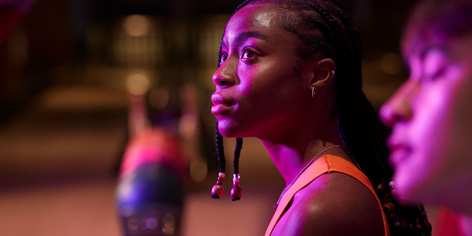 Person with braided hair in an orange top under purple lighting, with a blurred background.