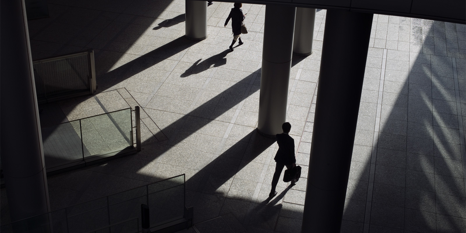 Two people walking through sunlit space with columns and long shadows.