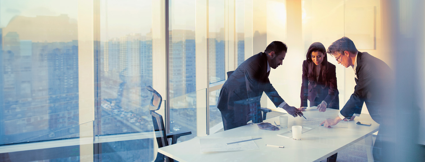 Four people in business attire gathered around a table in a modern office with city views.