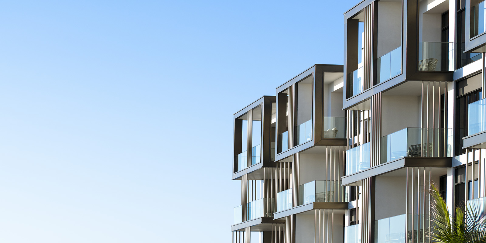 Modern apartment building with large windows and glass balconies, set against a clear blue sky.