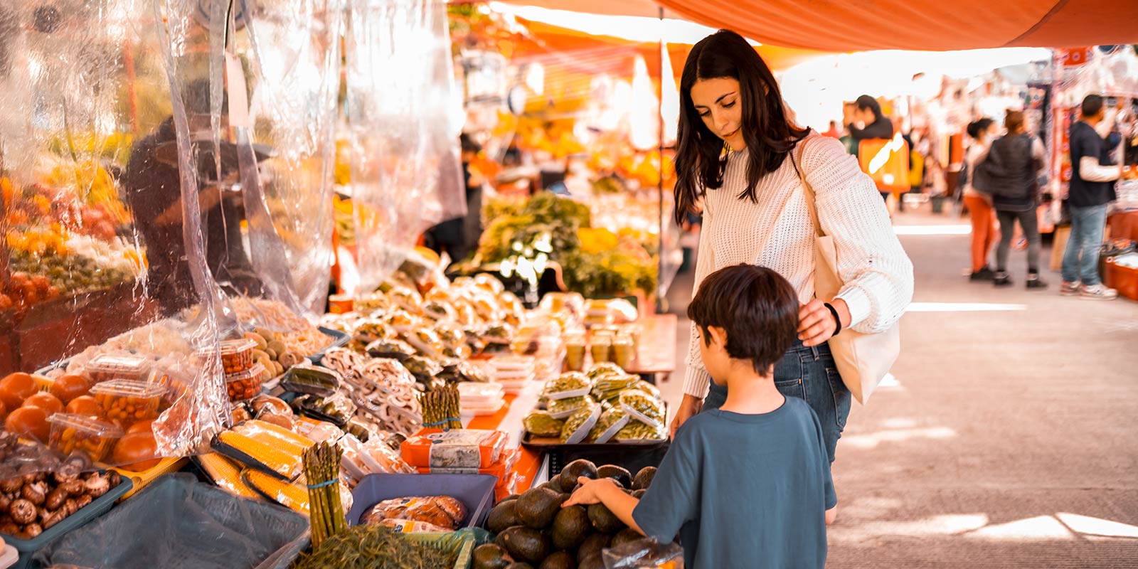 Outdoor market with orange canopies, neatly arranged produce, and a woman with a child near a stall of avocados.