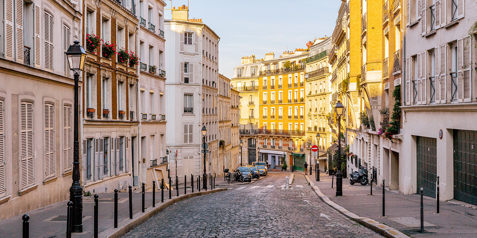 Cobblestone street with flowered balconies, parked vehicles, and warm lighting.