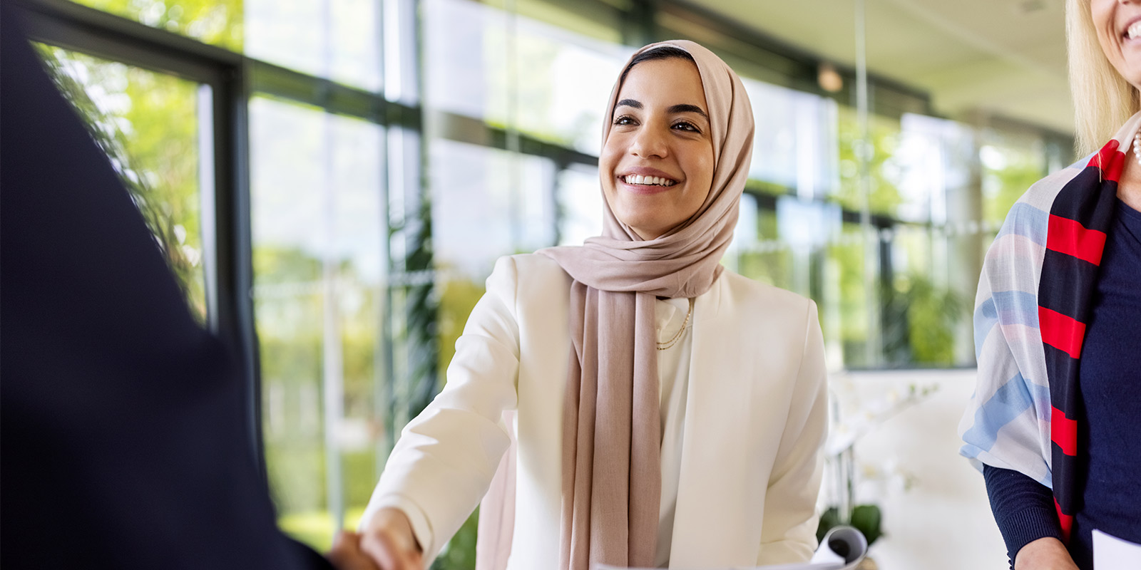 Three people indoors, with one in a hijab and white blazer offering a handshake.
